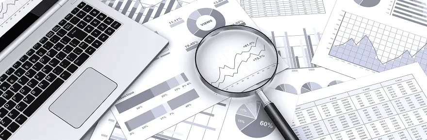 ノートパソコンとグラフ資料の上に置かれた虫眼鏡で、データ分析や統計資料を確認する様子